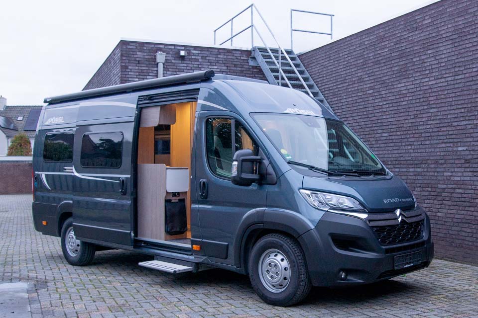 Een Pössl Roadcruiser buscamper met lengtebedden gezien vanaf de buitenzijde bij het vallen van de avond met de schuifdeur open en de verlichting van het interieur aan.